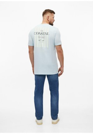 Homme portant un t-shirt bleu clair avec l'inscription « Coastal » dans le dos, jean bleu et baskets blanches, debout de dos sur fond blanc.