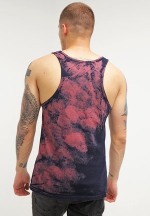 Mężczyzna z tatuażami na obu ramionach noszący bezrękawnik tie-dye w ciemnoniebieskim i czerwonym kolorze, zwrócony tyłem na jednolitym tle.