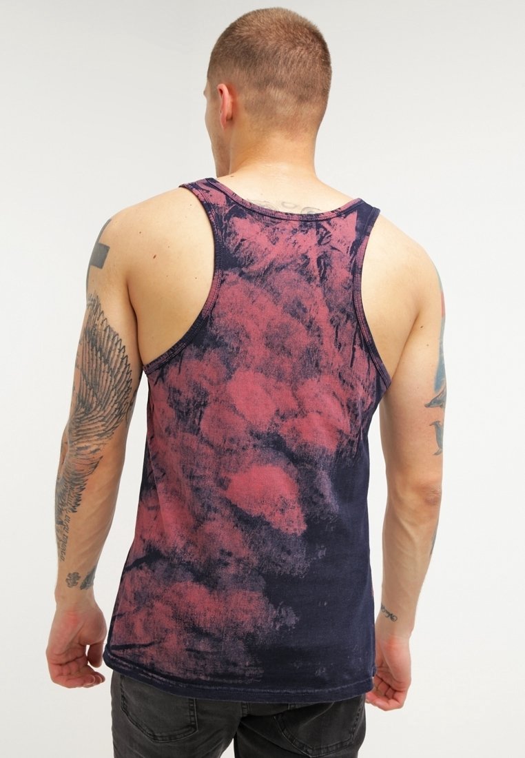 Homme avec des tatouages sur les deux bras, portant un débardeur tie-dye sans manches bleu foncé et rouge, tourné de dos sur un fond uni.