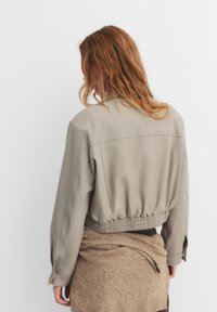 Afskåret, beige bomberjakke med elastisk talje og løse ærmer. Blødt, tekstureret stof. Båret over et brunt strikket plagg.