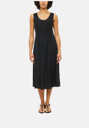 Woman wearing a sleeveless black midi dress with a scoop neckline and black T-strap flat sandals against a plain background.