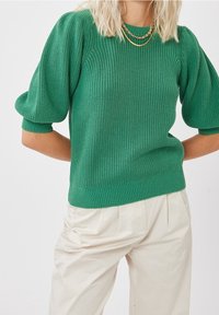 Femme portant un pull en maille vert à manches bouffantes et un pantalon plissé crème taille haute, accessoirisée avec des colliers en chaînes dorées superposés.