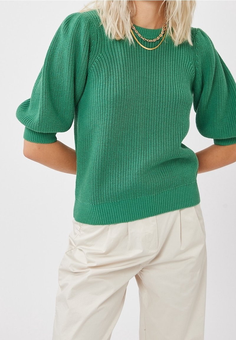 Femme portant un pull en maille vert à manches bouffantes et un pantalon plissé crème taille haute, accessoirisée avec des colliers en chaînes dorées superposés.