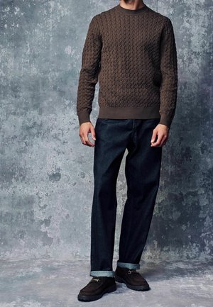 Pull en tricot marron avec un motif texturé, associé à un jean bleu foncé et des chaussures à lacets marron. Le pull présente un col rond et des poignets côtelés.