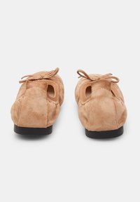Beige suede ballet flats with a flexible black sole, bow detail on top, and circular cutouts on the sides for breathability.
