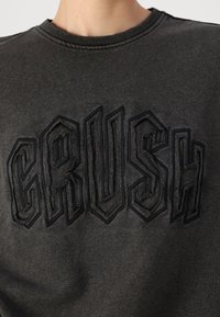 Felpa grigia scura con testo ricamato in rilievo nero "CRUSH". Il tessuto ha una texture liscia e un classico scollo rotondo.
