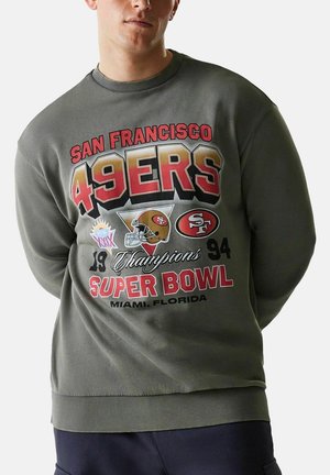 Mann trägt einen olivgrünen Pullover mit dem Logo der San Francisco 49ers Super Bowl 1994 Champions und einer Grafik eines Footballhelms.