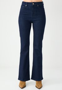 Jean évasé bleu foncé taille haute en denim, avec poches avant, coutures discrètes et légère texture. Accentué par des éléments métalliques.