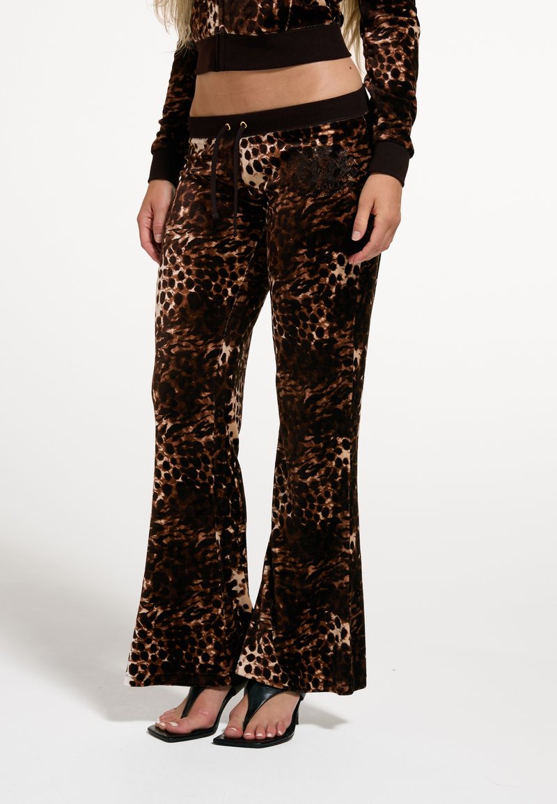 Γυναίκα φορά καφέ και μαύρο βελούδινο παντελόνι με animal print και μαύρα πέδιλα με τακούνι και ανοιχτά δάχτυλα, στέκεται μπροστά σε απλό λευκό φόντο.