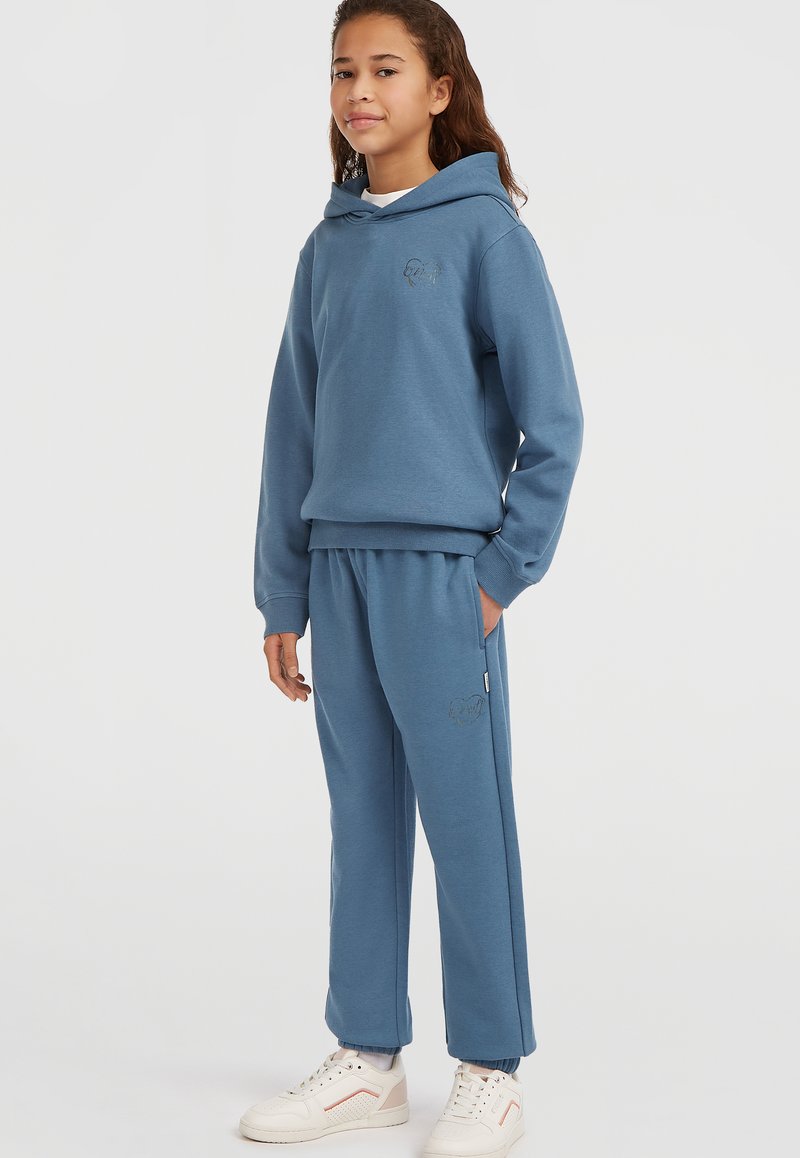 Sudadera azul con un bolsillo frontal y un logotipo bordado, acompañada de pantalones de chándal azul a juego. La tela parece suave, adecuada para llevar en ocasiones informales.