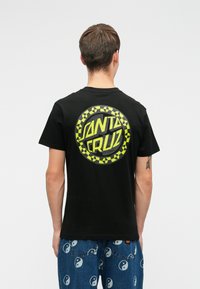 Άνδρας που φοράει μαύρο T-shirt Santa Cruz με κίτρινο λογότυπο και μπλε τζιν με λευκά σύμβολα γιν-γιάνγκ, στέκεται με την πλάτη στραμμένη προς την κάμερα σε απλό φόντο.
