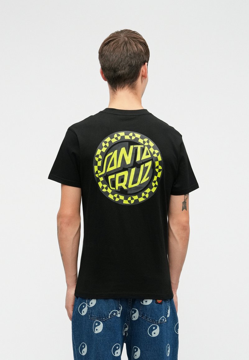 Άνδρας που φοράει μαύρο T-shirt Santa Cruz με κίτρινο λογότυπο και μπλε τζιν με λευκά σύμβολα γιν-γιάνγκ, στέκεται με την πλάτη στραμμένη προς την κάμερα σε απλό φόντο.
