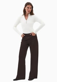 Mujer vestida con una camiseta blanca de manga larga y cuello con cremallera, y pantalones marrones de pierna ancha y tiro alto, con las manos en los bolsillos.