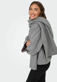 Grå ullblandad omhängd kappa med en stor huva, oversized ärmar och inre rosa foder. Modellen står i en trekvart vy mot en enkel bakgrund.