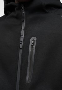 Μαύρο φούτερ με κουκούλα και λεία υφή, με κάθετη τσέπη με φερμουάρ και αντανακλαστικές λεπτομέρειες. Η λέξη "CORE" τυπωμένη μαύρη κατά μήκος της γραμμής του φερμουάρ.