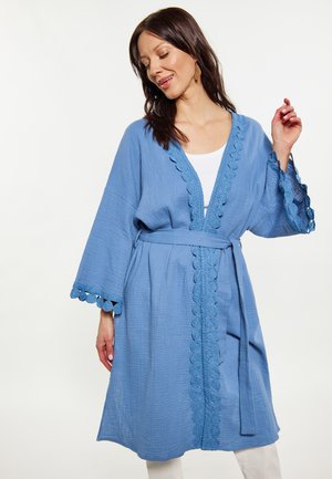 Woman wearing a knee-length blue textured robe with scalloped lace trim and a waist tie, paired with white clothing underneath.
