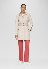 Lichtbeige trenchcoat met een ceintuur in de taille, gouden knopen en twee voorzakken. In combinatie met koraalkleurige broek en witte platform sneakers.