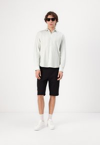 Chemise à manches longues vert clair avec des rayures verticales, short noir et baskets blanches, portée avec des lunettes de soleil noires. Figure debout.
