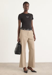 T-shirt nera aderente con testo bianco, pantaloni a gamba larga beige con righe, cintura nera, borsa nera con design intrecciato e tacchi a punta neri.