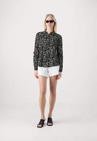Black button-up shirt with white abstract pattern, paired with white frayed hem shorts and black open-toe mules. Model poses against a plain background.