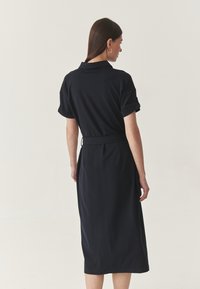 Rochie bleumarin cu curea, cu mâneci scurte răsucite, un guler clasic și o siluetă elegantă, realizată dintr-un material moale, texturat.