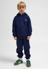 Mörkblå sweatshirt och joggers-set i mjukt tyg, med en stor framficka, ribbade ärmslut och gröna sneakers med vita detaljer.
