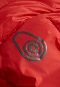 Rød dunjakke med et fremtrædende sort logo med et cirkulært design og koncentriske ringe, der viser en glat tekstur.