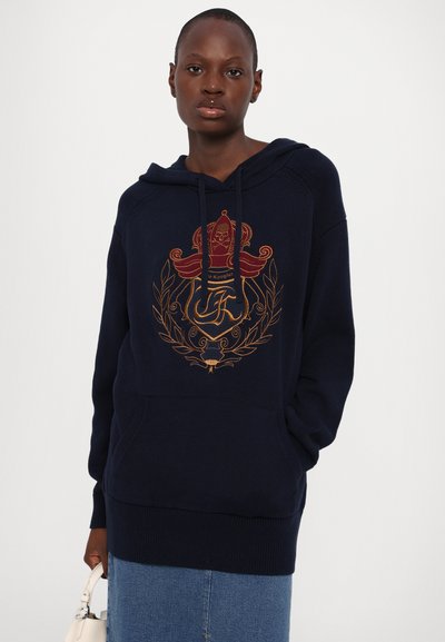 Tumesinine kapuutsiga särk, millel on punane ja kuldne tikandiga emblem, millel on kroon ja laureli muster. Materjal näib olevat kudum ja esiküljel on tasku.