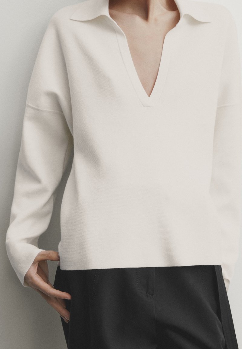 Sweater en tricot blanc avec un large col en V et un col, présentant des épaules tombantes et une coupe décontractée. Associe à un pantalon noir.