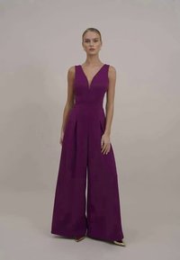Lilla ärmlös jumpsuit med djupt V-hals, åtsittande bodice, vida plisserade ben och mjukt tyg. Modellen poserar framför en neutral bakgrund.