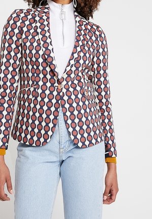 Frau trägt ein gemustertes Blazer mit geometrischem Design, weißem Reißverschluss-Top und hellblauen Jeans vor einfarbigem Hintergrund.