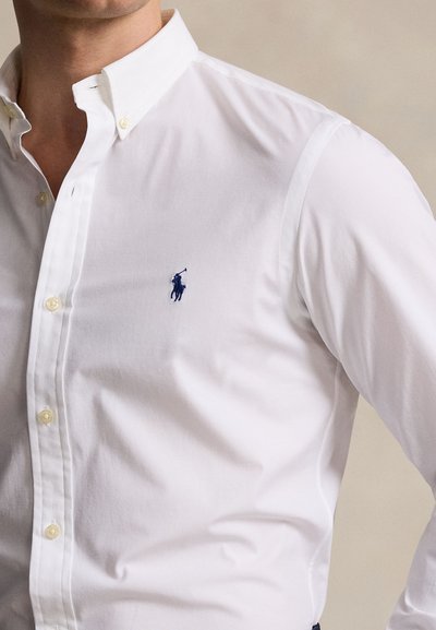 Muž nosiaci bielu košeľu na gombíky s golierom a malým tmavomodrým vyšívaným logom hráča póla na hrudi.