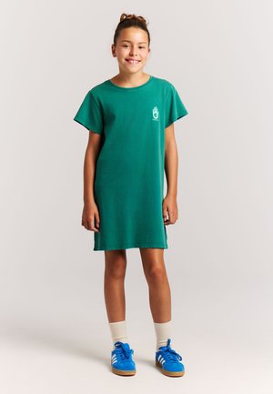 Groene katoenen t-shirtjurk met korte mouwen en een losse pasvorm. Voorzien van een kleine afbeelding op de borst. Gecompleteerd met blauwe sportschoenen.