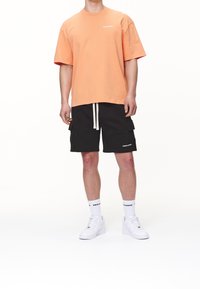 Hombre con camiseta oversized de color melocotón, pantalones cortos cargo negros con cordón blanco, calcetines con logo blancos y zapatillas blancas, de pie sobre un fondo blanco.