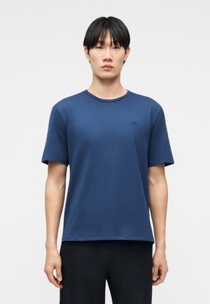 Bărbat tânăr cu păr negru drept, purtând un tricou simplu albastru cu mâneci scurte și pantaloni negri, stând în fața unui fundal simplu deschis la culoare.