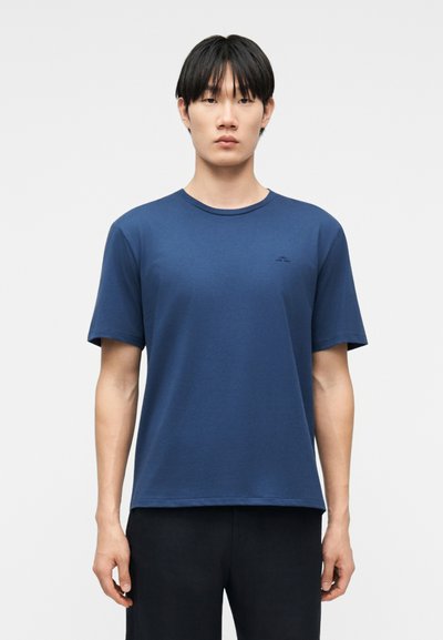 Giovane con capelli neri lisci che indossa una maglietta blu a maniche corte semplice e pantaloni neri, in piedi contro uno sfondo chiaro e uniforme.