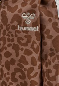 Tissu brun avec un motif imprimé léopard ; présente un logo "hummel" brodé en blanc. Texture lisse avec une finition légèrement brillante.