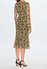 Kleid mit langen Ärmeln aus transparentem Stoff mit braunem Grund und bunten, wirbelnden Blattmustern. Es verfügt über einen ausgestellten Saum und wird mit schwarzen High Heels kombiniert.