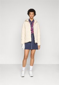 Veste en crème rembourrée avec capuche, portée sur un haut sombre à motifs et une jupe bleu marine. Des chaussures de sport blanches et un gant blanc accentuent la tenue.