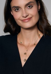 Złoty naszyjnik z łańcuchem, ozdobiony dwoma zawieszkami w kształcie liści oraz akcentami w postaci zielonych kamieni szlachetnych, noszony z czarną bluzką, podkreślając elegancki design.