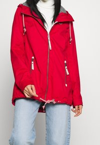 Červená, voděodolná bunda s šedou kapucí. Obsahuje dvě boční kapsy se zipem, nastavitelné stahování na lemu a kontrastní kovové detaily.