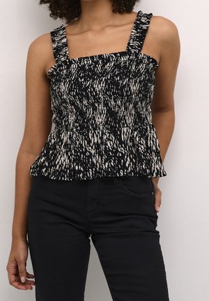 Kobieta w czarno-białym, bezrękawnym topie z falbanami, wzorzystym, zestawionym z czarnymi spodniami, z ręką w kieszeni, na jednolitym tle.