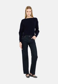 Maglione a maglia navy con scollo ampio abbinato a jeans blu scuro a gamba dritta. Il modello indossa delle ballerine nere contro uno sfondo neutro.