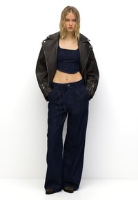 Temno modra skrajšana majica in široke temno modre jeans hlače v kombinaciji z rjavim usnjenim biker jakno, ki ima srebrno okovje in zadrge.