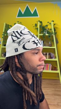 Berretto in maglia bianco con scritta nera. Presenta una vestibilità aderente e un'etichetta cucita. Sullo sfondo muro giallo e scaffali verdi.