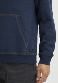 Marinast bluza iz mehkega bombažnega materiala, z sprednjim žepom, kontrastnim šivanjem in etiketo na boku.