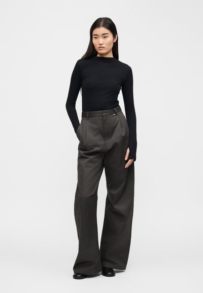 Γυναίκα που στέκεται με τα χέρια στις τσέπες του παντελονιού, φορώντας μαύρο μακρυμάνικο τοπ και φαρδιά σκούρα γκρι παντελόνια, μπροστά σε απλό φόντο.