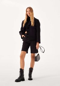 Fekete, rövidített fazonú zakó gombos elejjel, hozzá illő, szabott fekete rövidnadrággal párosítva. A modell csipkefűzős fekete bakancsot visel, és egy kis fekete kézitáskát tart a kezében.