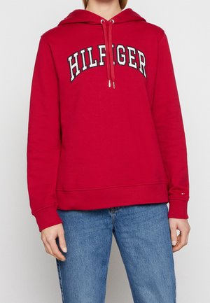 Osoba w czerwonej bluzie z kapturem z napisem "HILFIGER" na piersi i niebieskich dżinsach, stojąca na tle jednolitego jasnego tła.