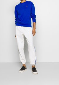 Blå sweatshirt med rund halsringning och liten logotyp, tillsammans med vita mjukisbyxor med blå sidor, och svarta sneakers.